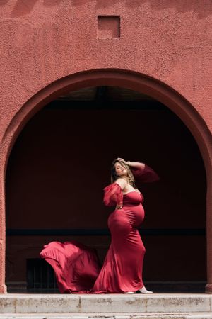 Dramatic maternity portrait of a woman in red gown posing within a terracotta archway.