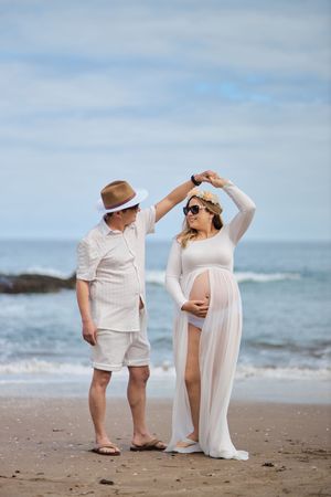 A couple in white outfits shares playful moments together on a sandy beach with ocean waves in the background.