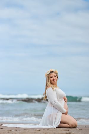 A person in a white dress and floral crown poses on a beach with waves crashing in the background.