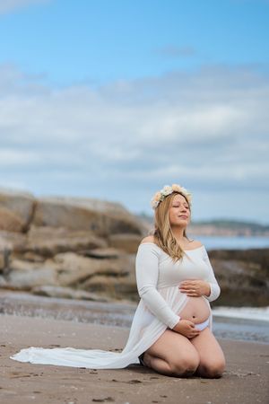 Expectant mother in white dress and floral crown poses serenely on beach with rocky coastline and cloudy sky backdrop.