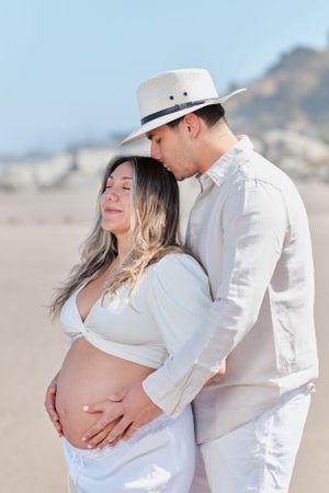 Intimate beach maternity photo with couple in white attire against mountain backdrop.