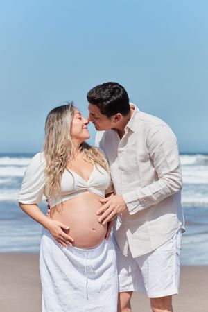 Romantic beach maternity photo with couple in white clothing by the ocean.