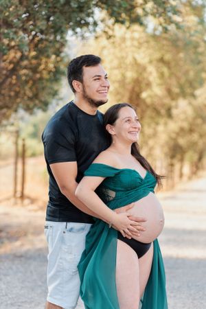 Maternity photo of couple in casual wear embracing on tree-lined path.