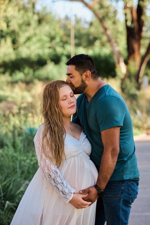 A couple embraces outdoors in a romantic maternity photoshoot with natural greenery in the background.
