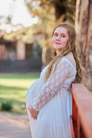Expectant mother in white lace maternity dress poses outdoors during golden hour sunset.