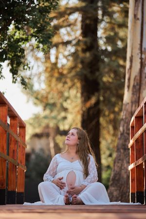 A person in a white lace dress sits thoughtfully on a wooden deck surrounded by autumn trees.