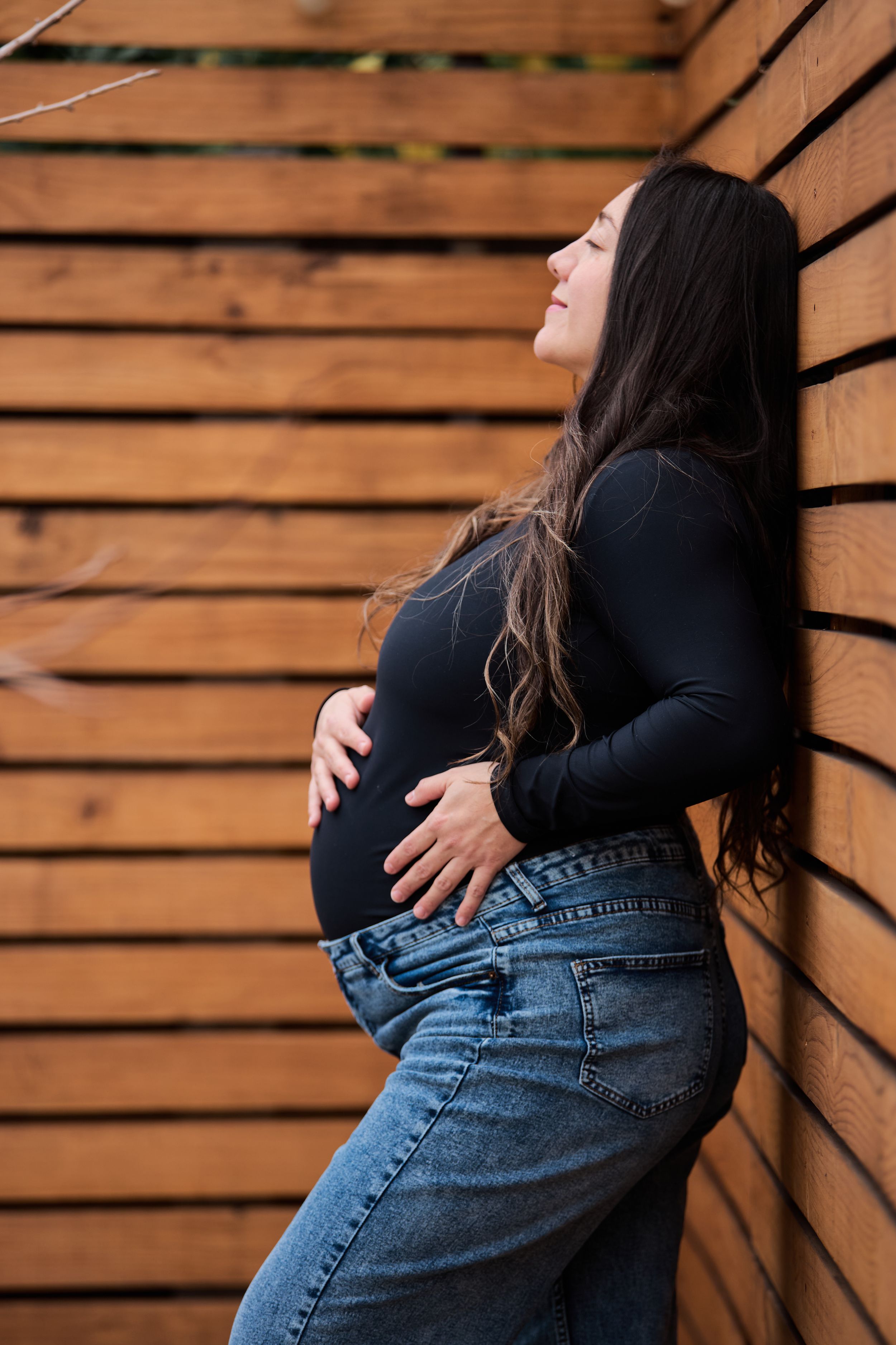 Maternity photo series showing pregnant woman in black top and jeans against wooden wall in natural lighting.