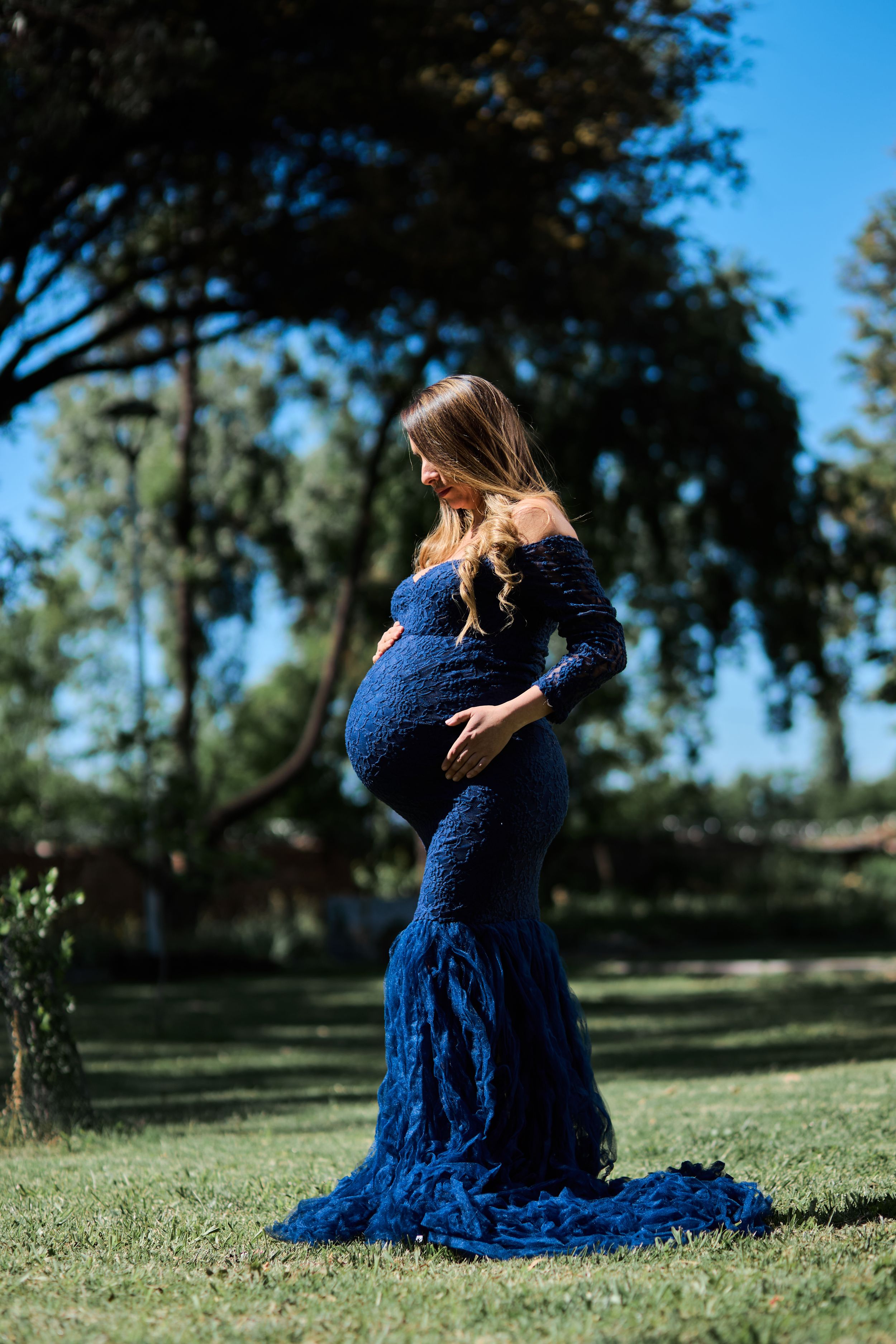 A figure in a flowing navy blue lace dress twirls gracefully on green grass in a sunlit outdoor setting.