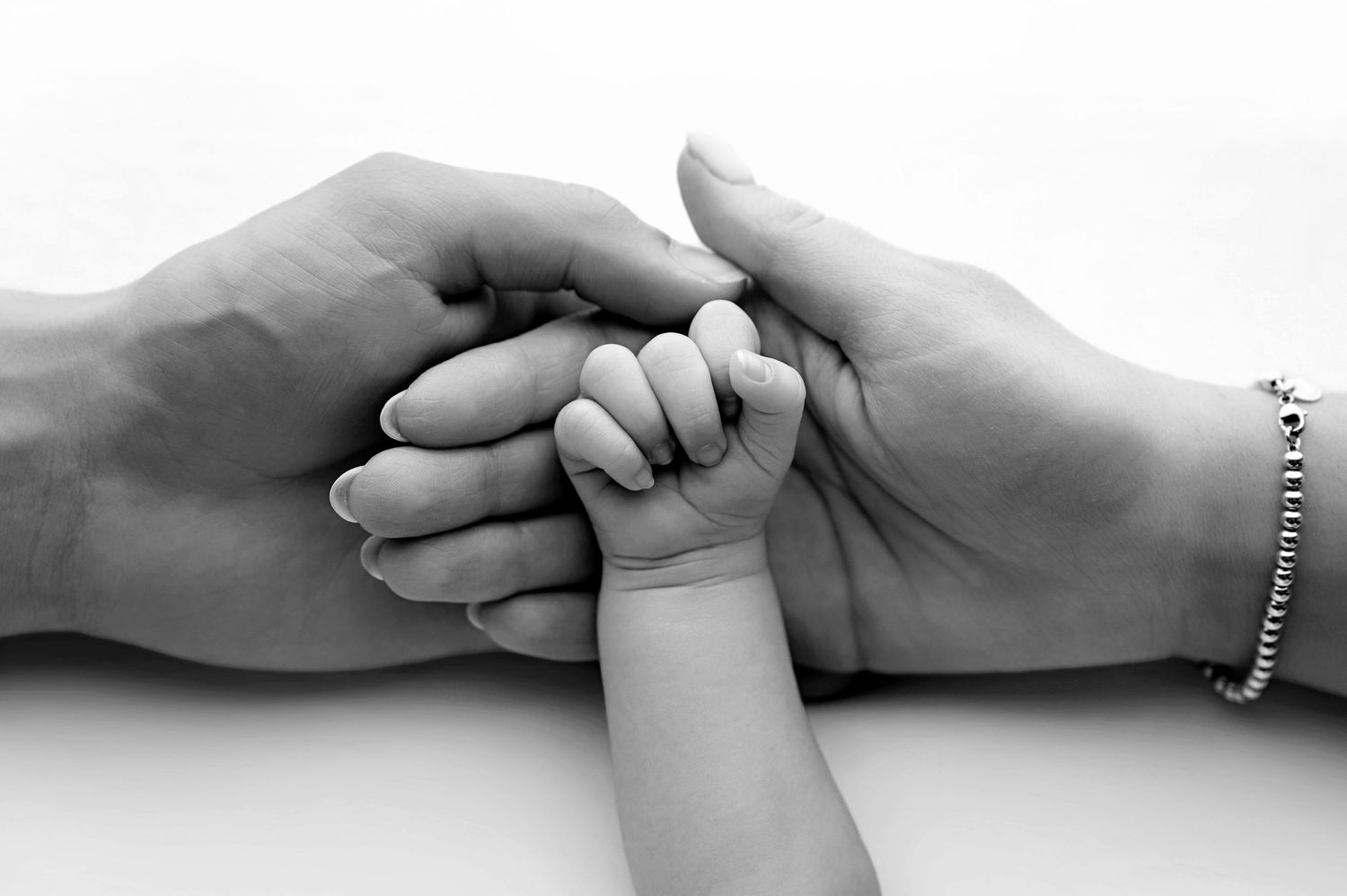 New parents in white shirts tenderly hold their sleeping newborn wrapped in a brown swaddle against a beige background.