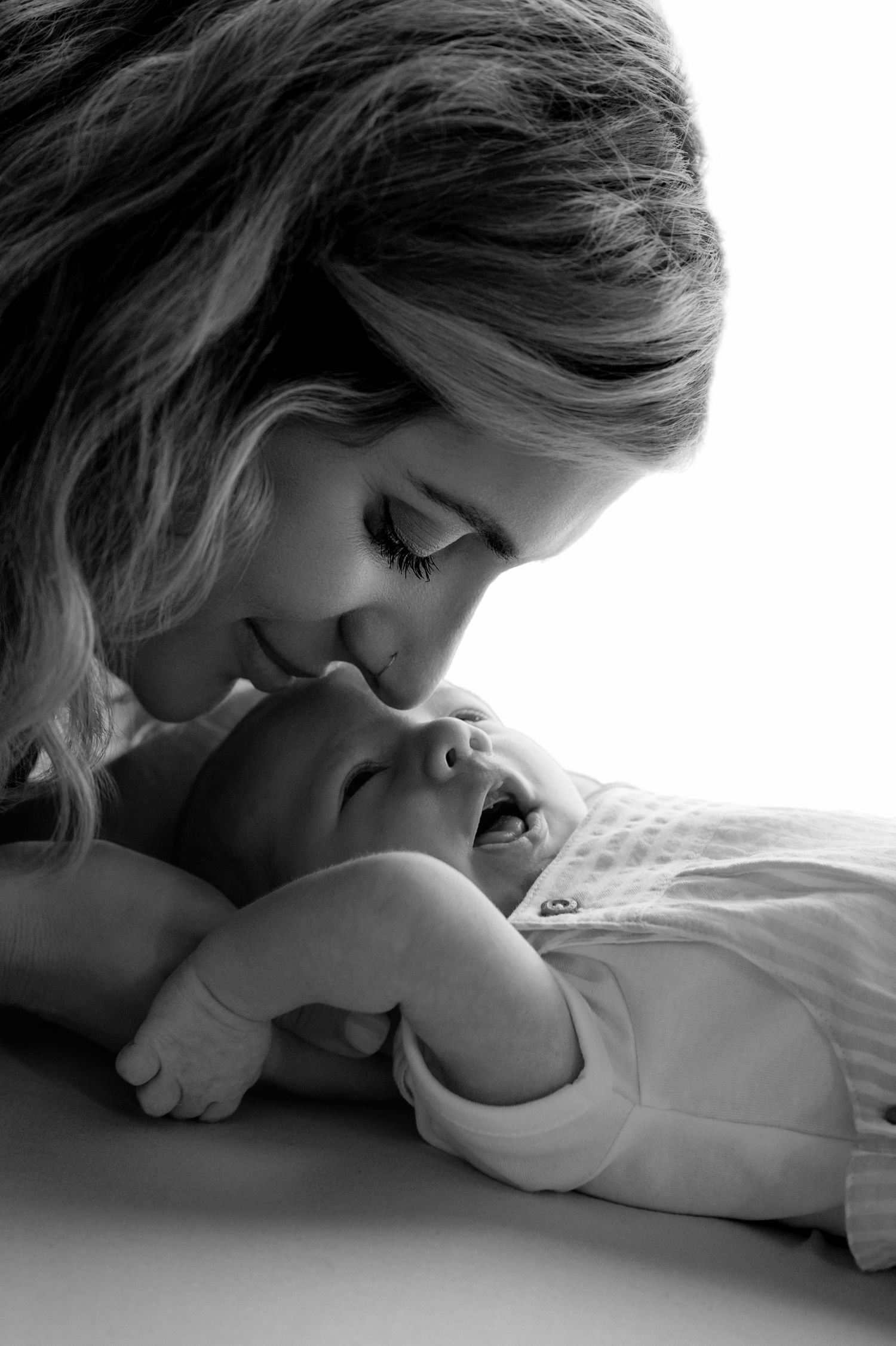 New parents in white shirts tenderly hold their sleeping newborn wrapped in a brown swaddle against a beige background.