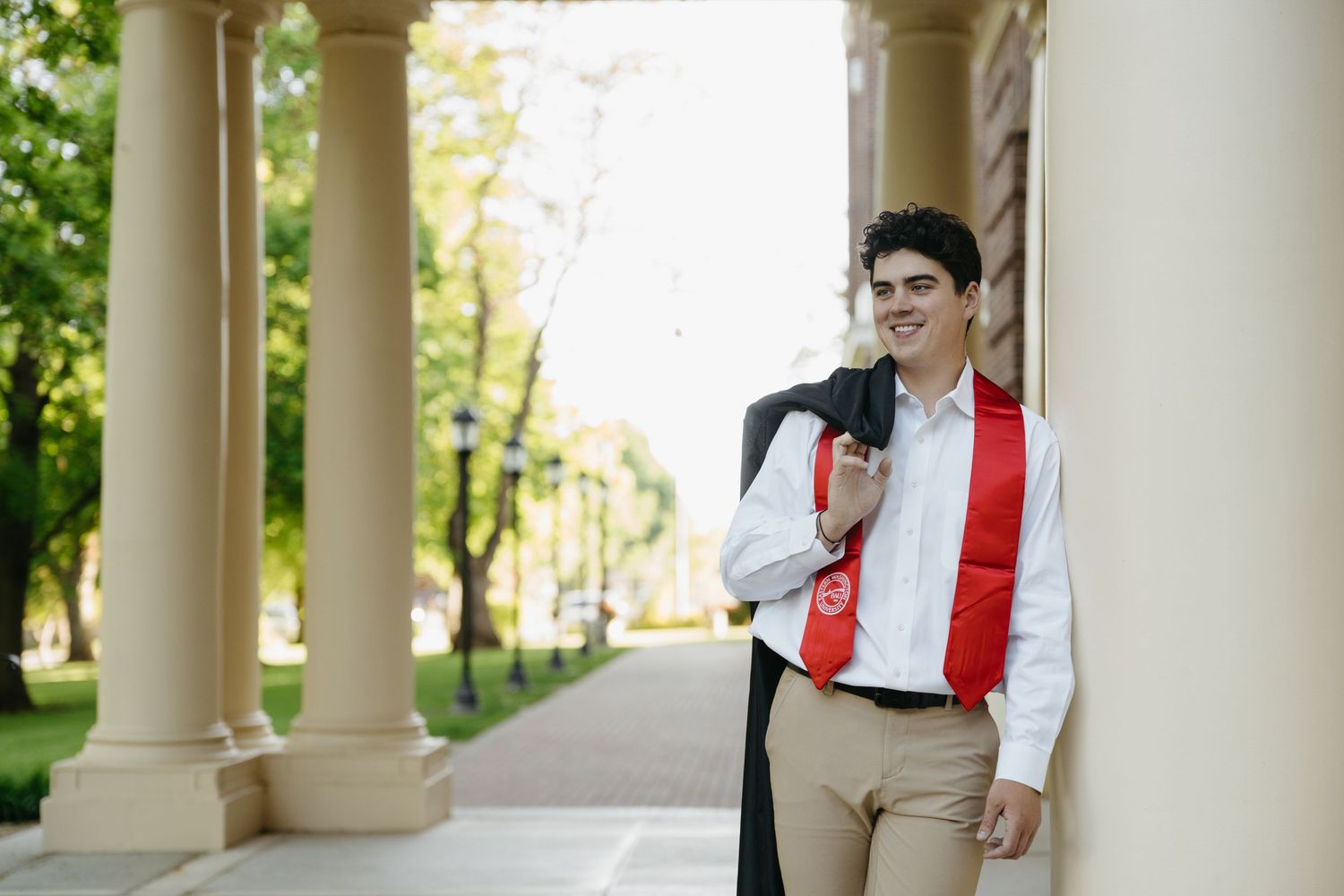 Young male graduate in white shirt and red sash holds cap and gown beside campus columns outdoors.