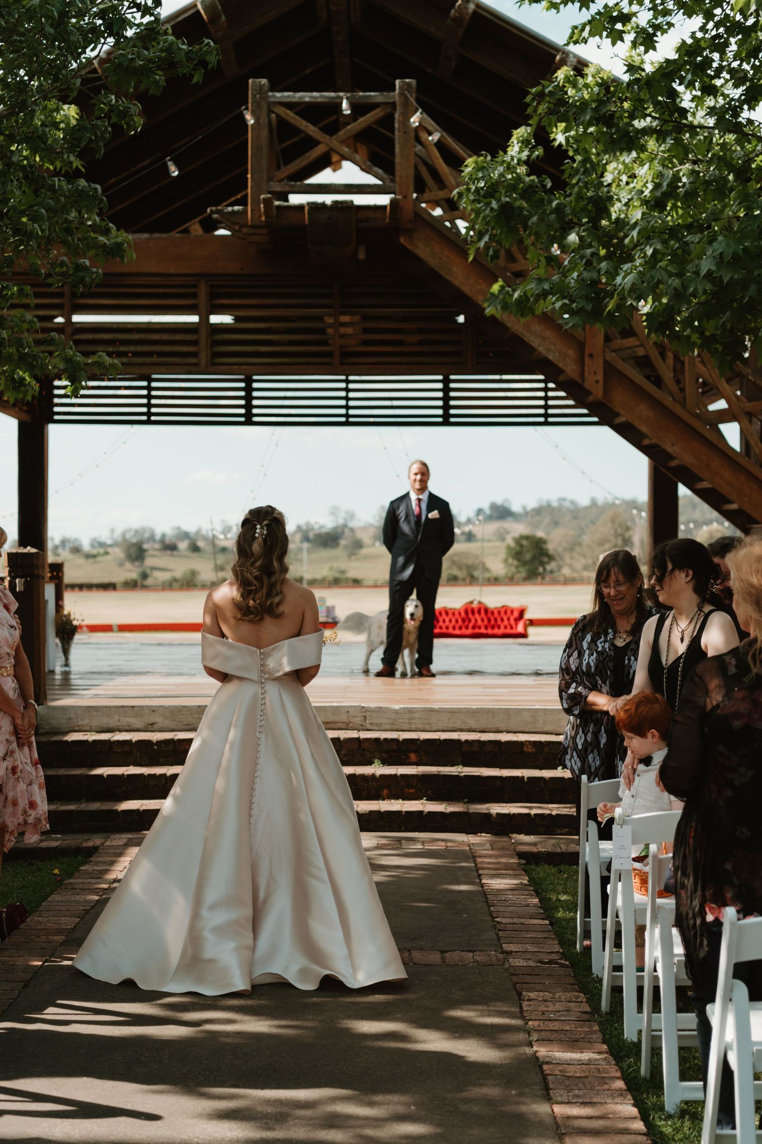 Bride walks down outdoor aisle toward groom under wooden pavilion at waterfront wedding ceremony.