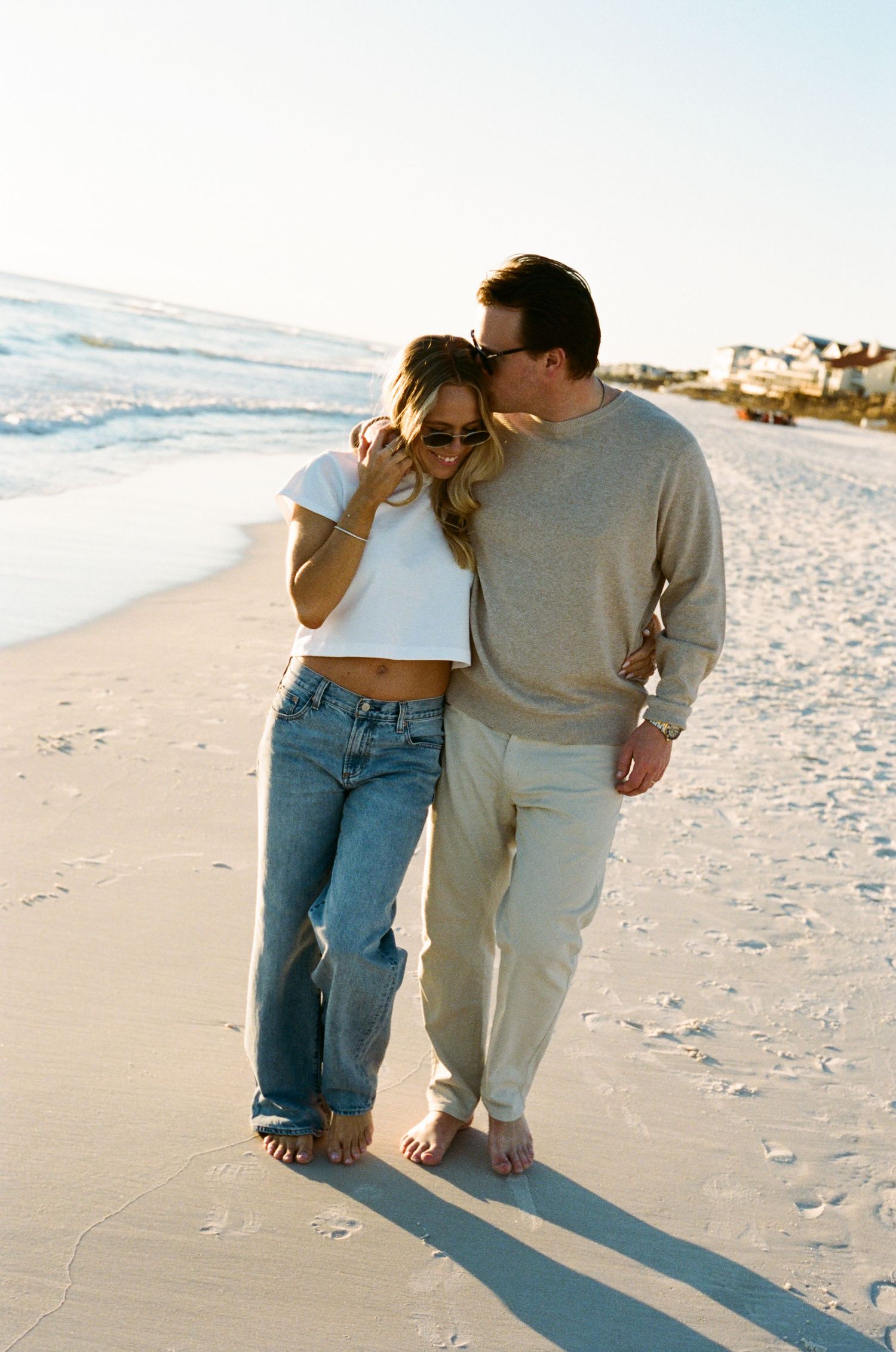 Couple walking barefoot on a sandy beach at sunset, woman in crop top and jeans, man in beige sweater.