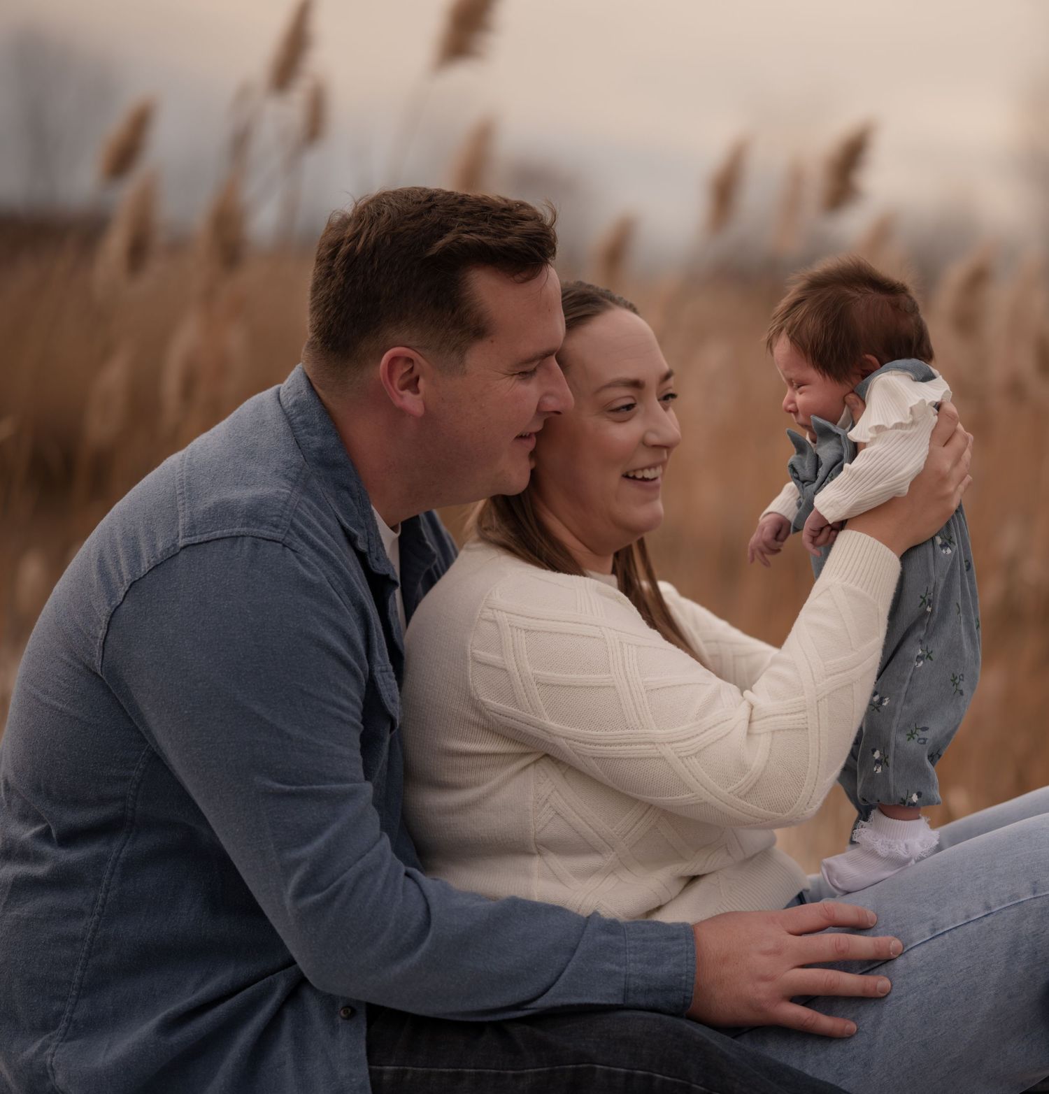 A happy couple sits outdoors in a field, smiling as they hold their newborn baby in soft natural light.