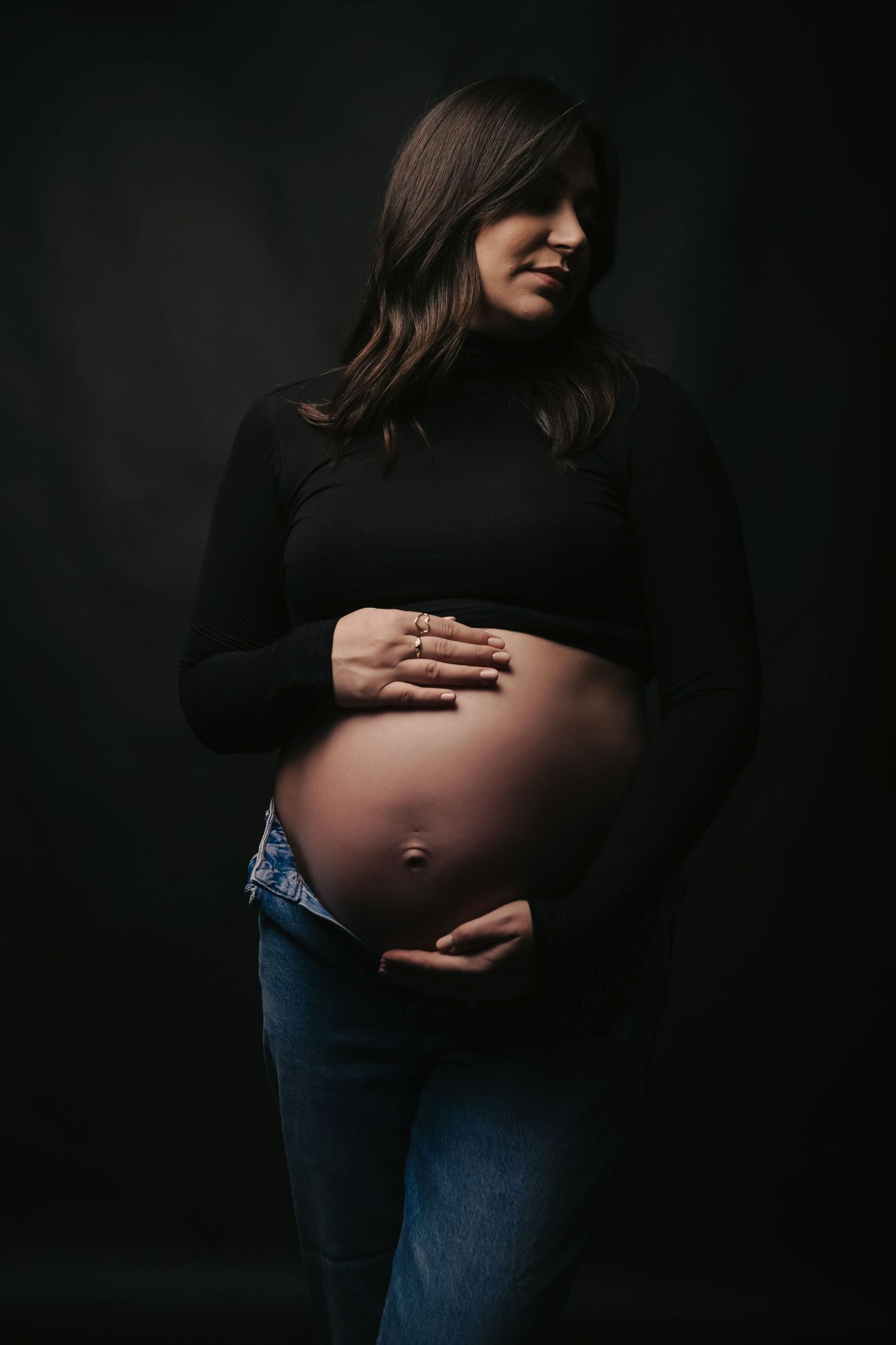 Moody maternity portrait of pregnant woman in black sweater and jeans against dark background.