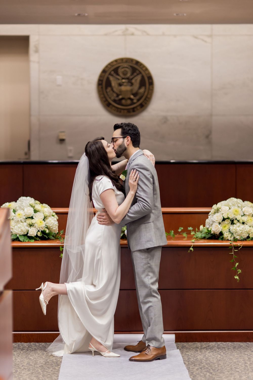 A couple in wedding attire share romantic kisses in a courthouse setting with floral arrangements in the background.