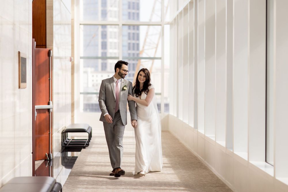 A couple walks together in an elegant white hallway with natural light streaming through tall windows.