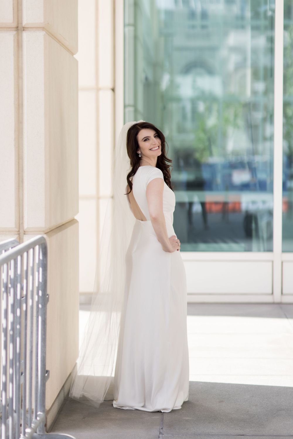 A bride in a flowing white dress poses gracefully along a bright, sunlit colonnade with multiple reflections.