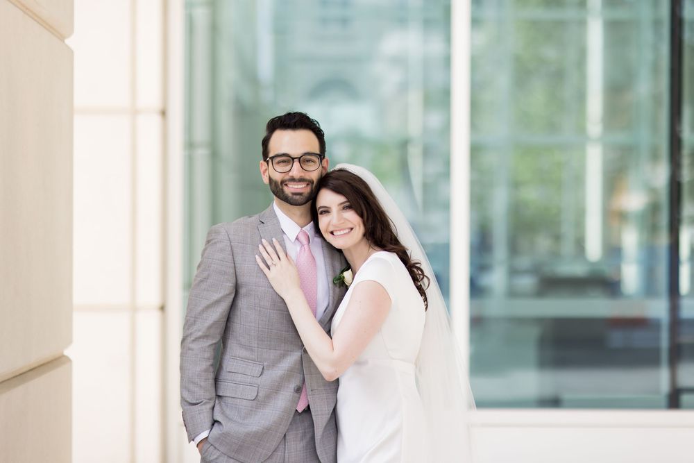 A couple in wedding attire share a romantic moment in front of a modern building with glass windows.