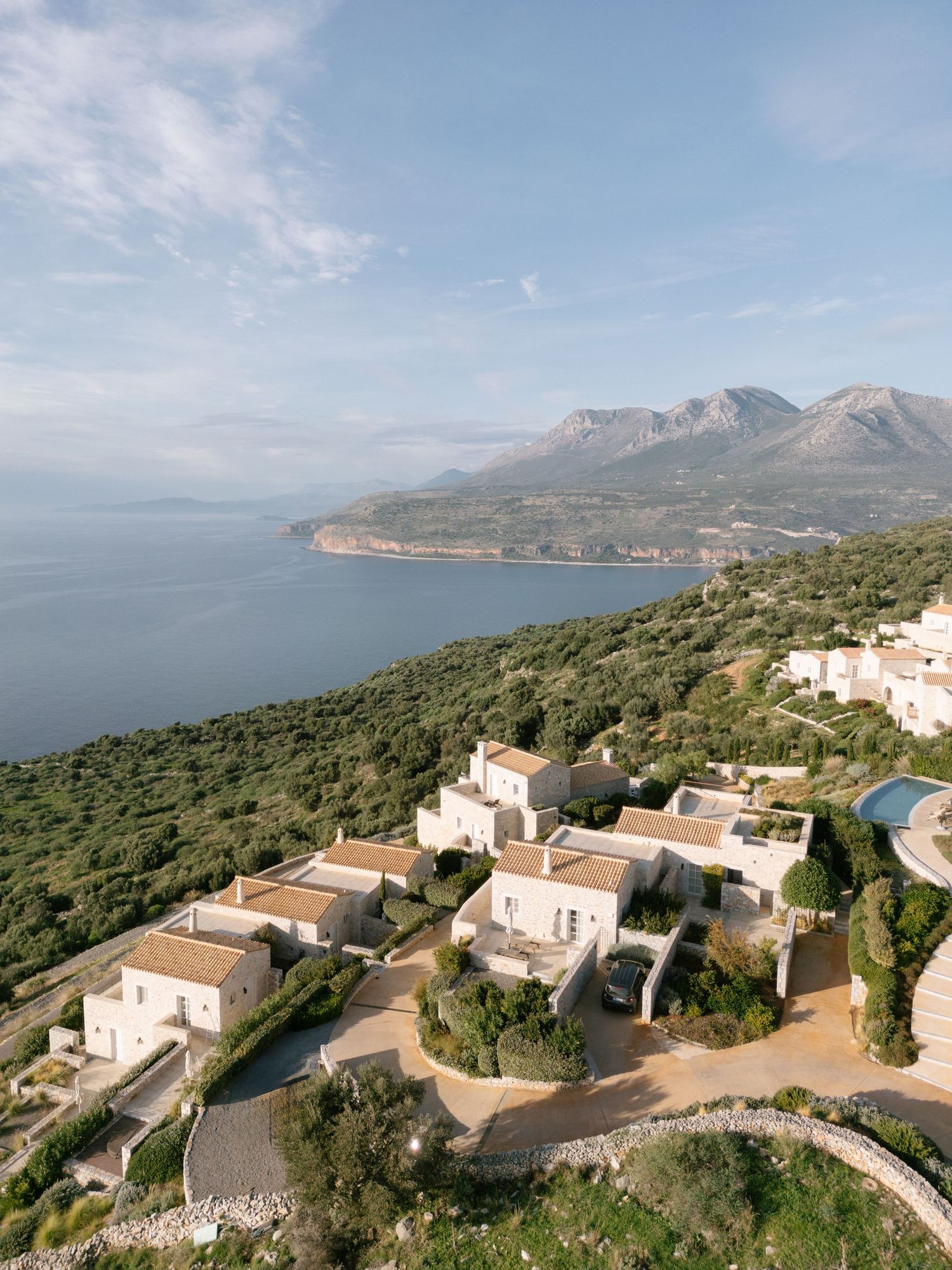 Panoramic coastal view of Mediterranean hillside villas overlooking blue waters and rugged mountains in the distance.