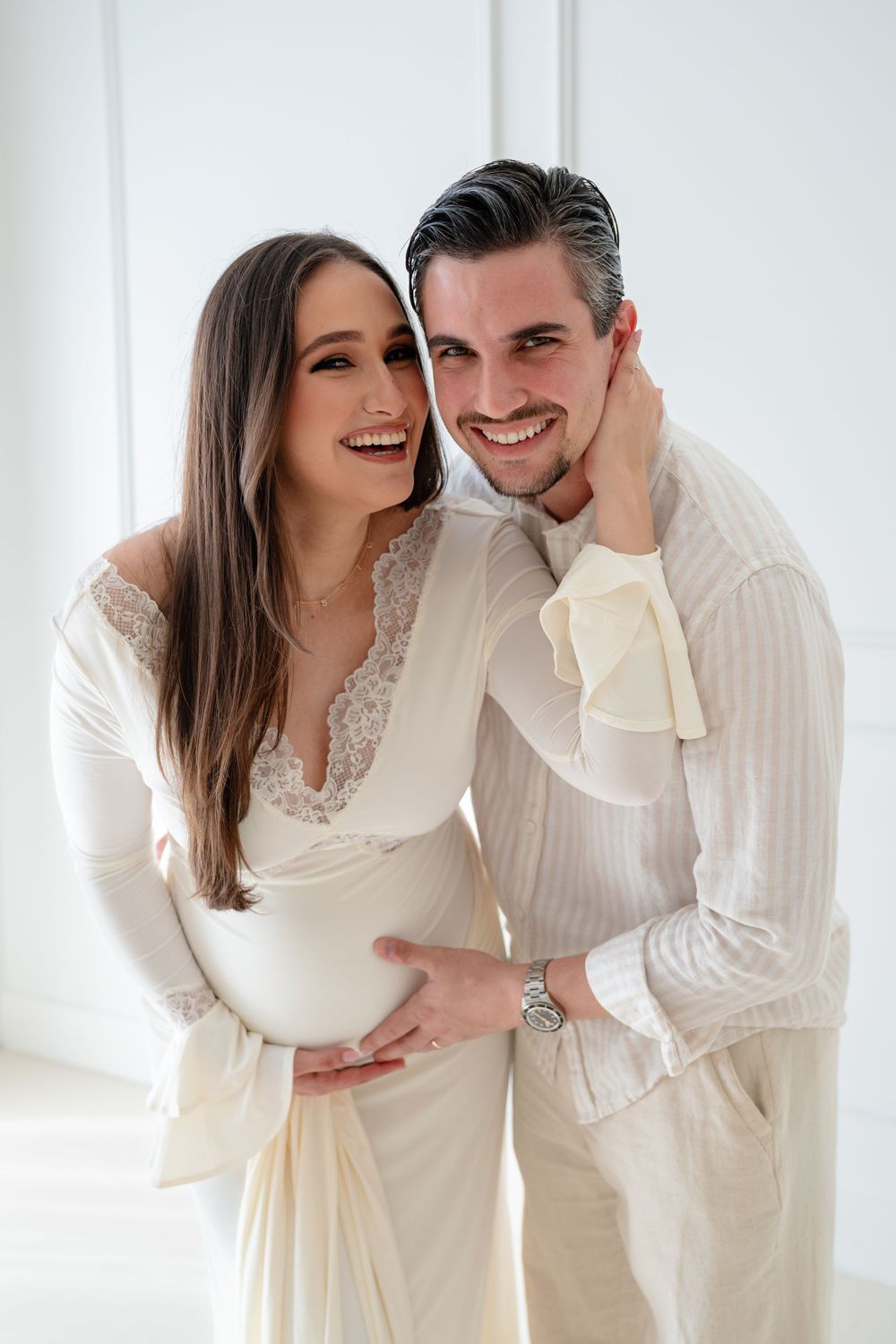 A couple in white outfits share intimate moments and laughter during a bright indoor photo session.