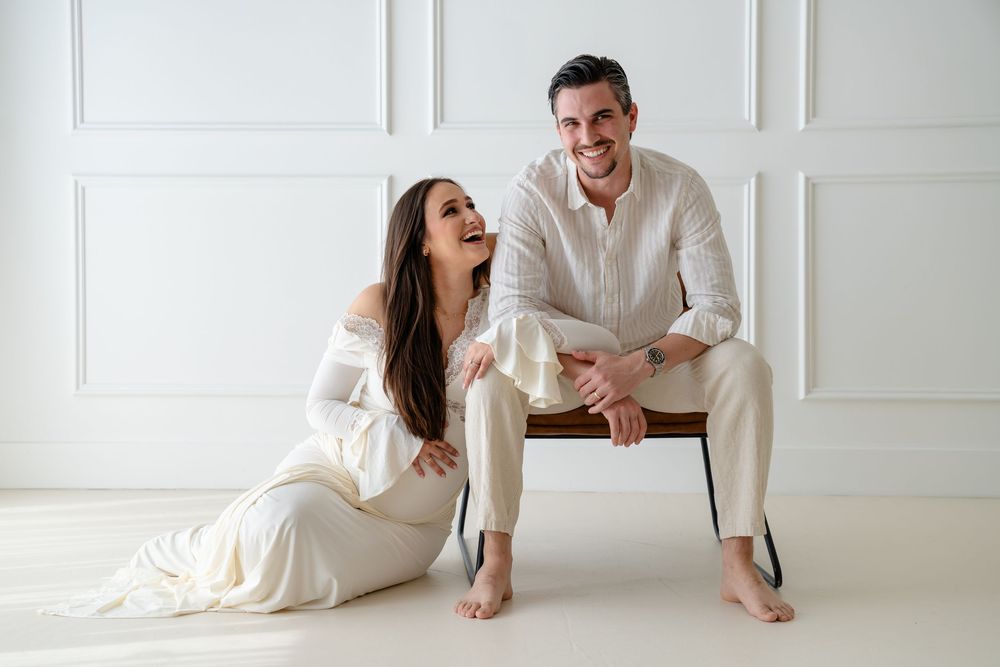 A couple in white clothing lies on a white floor in a bright minimalist setting during a photoshoot.