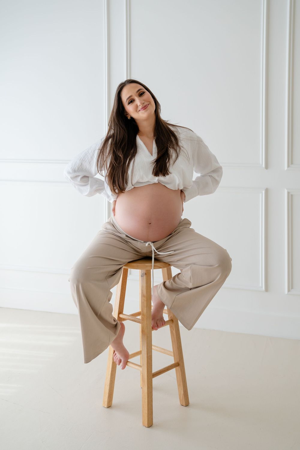 A maternity photoshoot series showing a pregnant woman in white and beige clothing posing on a wooden stool.