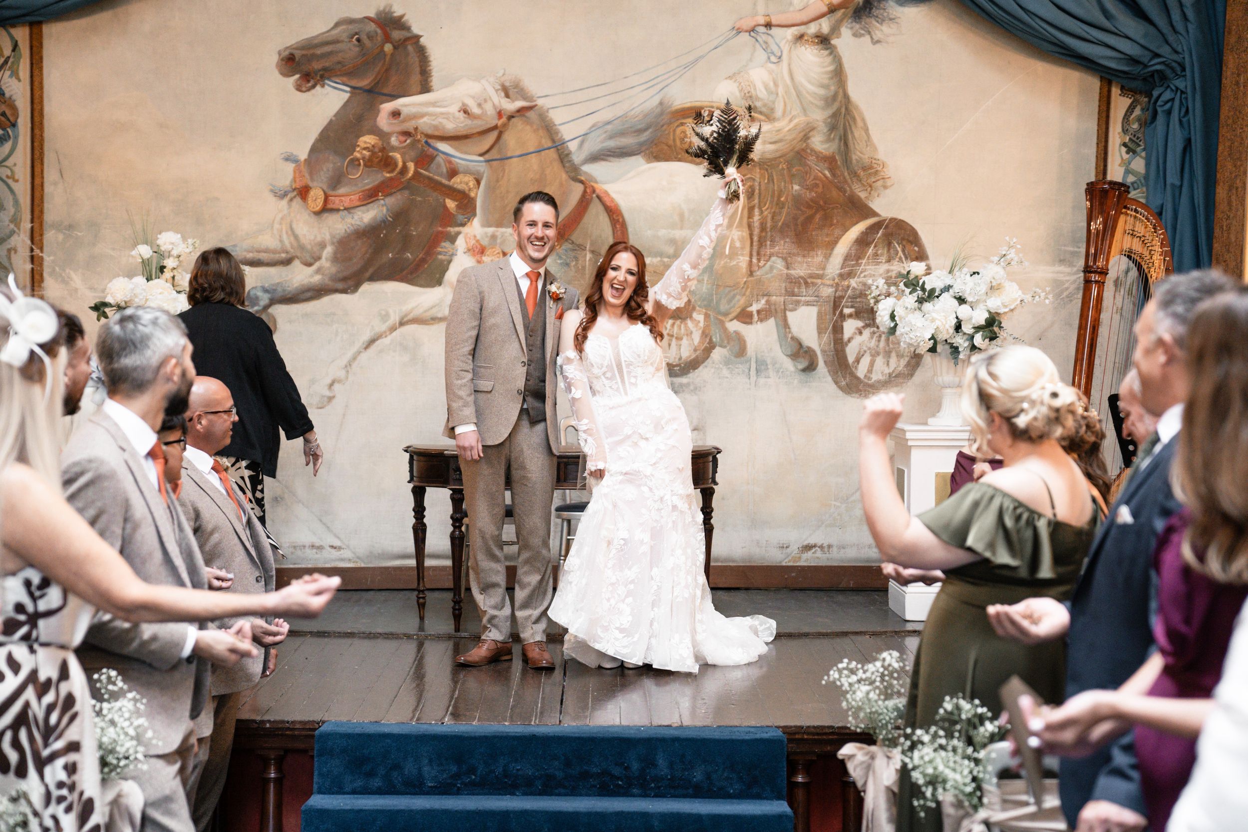 A wedding couple and guests dance together in a rustic barn venue decorated with string lights and murals on the walls.