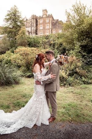 A romantic wedding couple poses in front of a historic stone building surrounded by lush gardens.