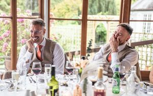 Wedding guests seated at long tables in a bright sunlit reception venue share joyful laughter during celebrations.