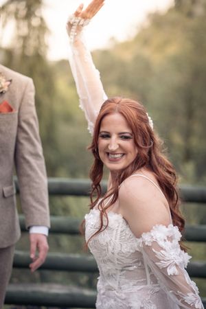 A bride in a white lace off-shoulder wedding dress smiles joyfully while standing outdoors.