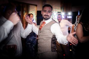 A wedding guest in a tan vest and orange tie dances energetically at an indoor reception.