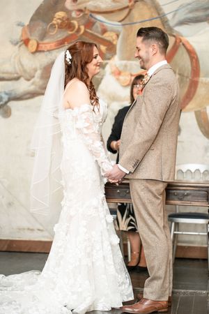 A wedding ceremony in front of a painted mural backdrop with the couple in white lace and tan suit.