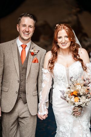 A bride in a lace gown and groom in grey suit with orange tie share candid moments during their wedding portraits.