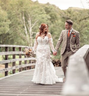 Newlywed couple walking along a rustic wooden bridge surrounded by autumn foliage during their wedding photoshoot.