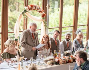 Wedding guests enjoy a celebratory meal at long tables in a sunlit rustic venue with wooden beams and windows.