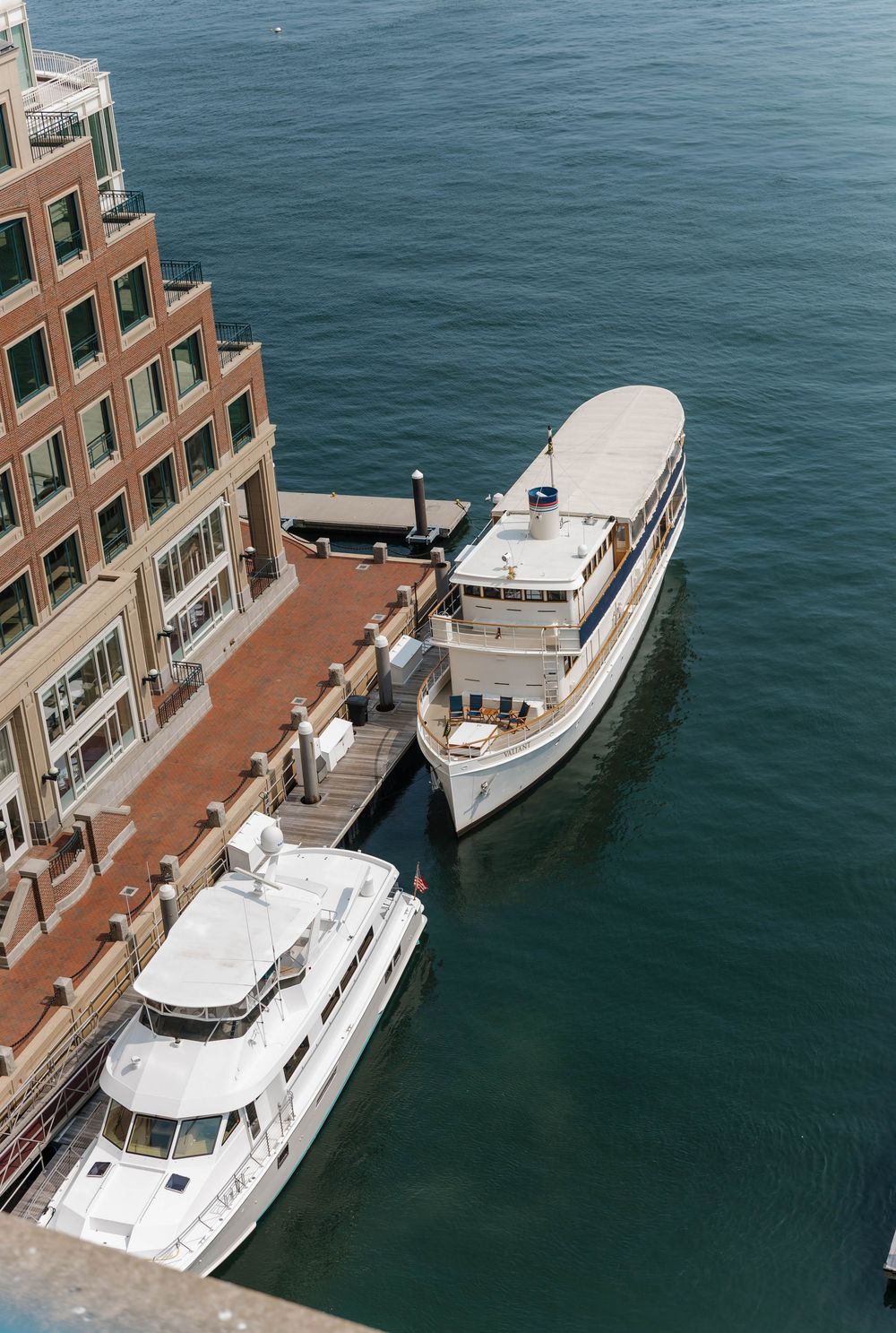 Aerial view of two luxury boats docked at a brick waterfront building on calm blue waters.