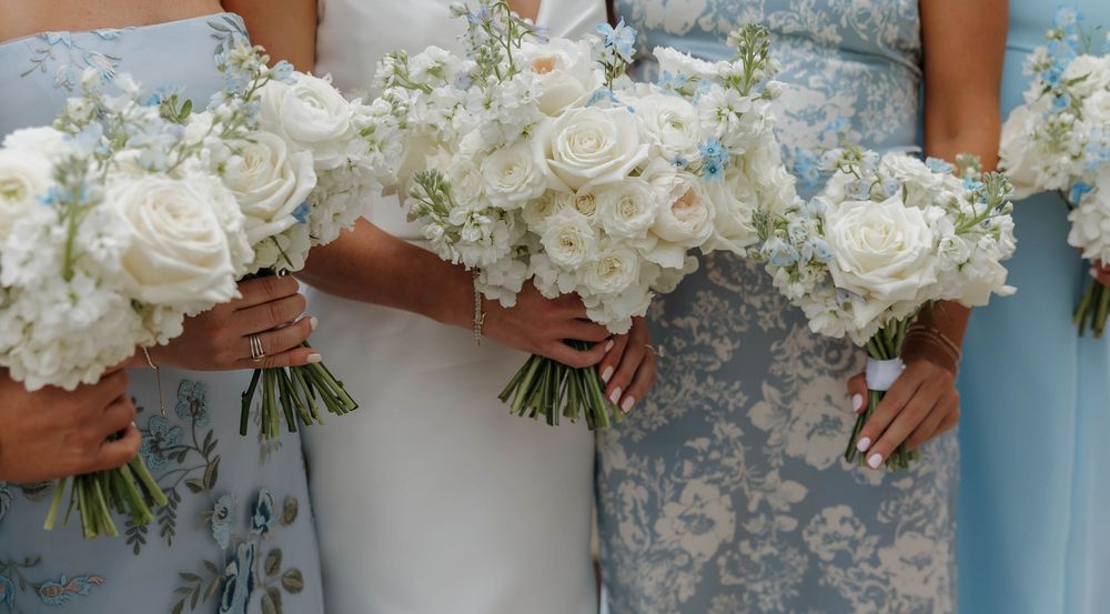 Three elegant white rose and baby's breath wedding bouquets held against light blue bridesmaid dresses.