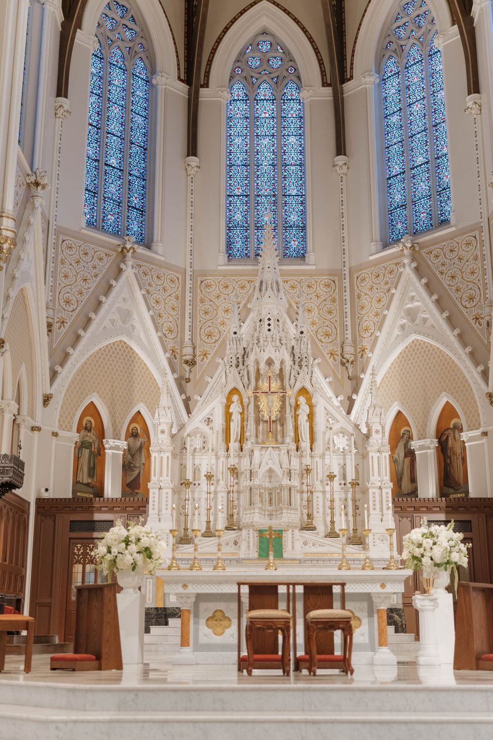 Ornate white Gothic church altar with tall stained glass windows and decorative floral arrangements in a cathedral sanctuary.