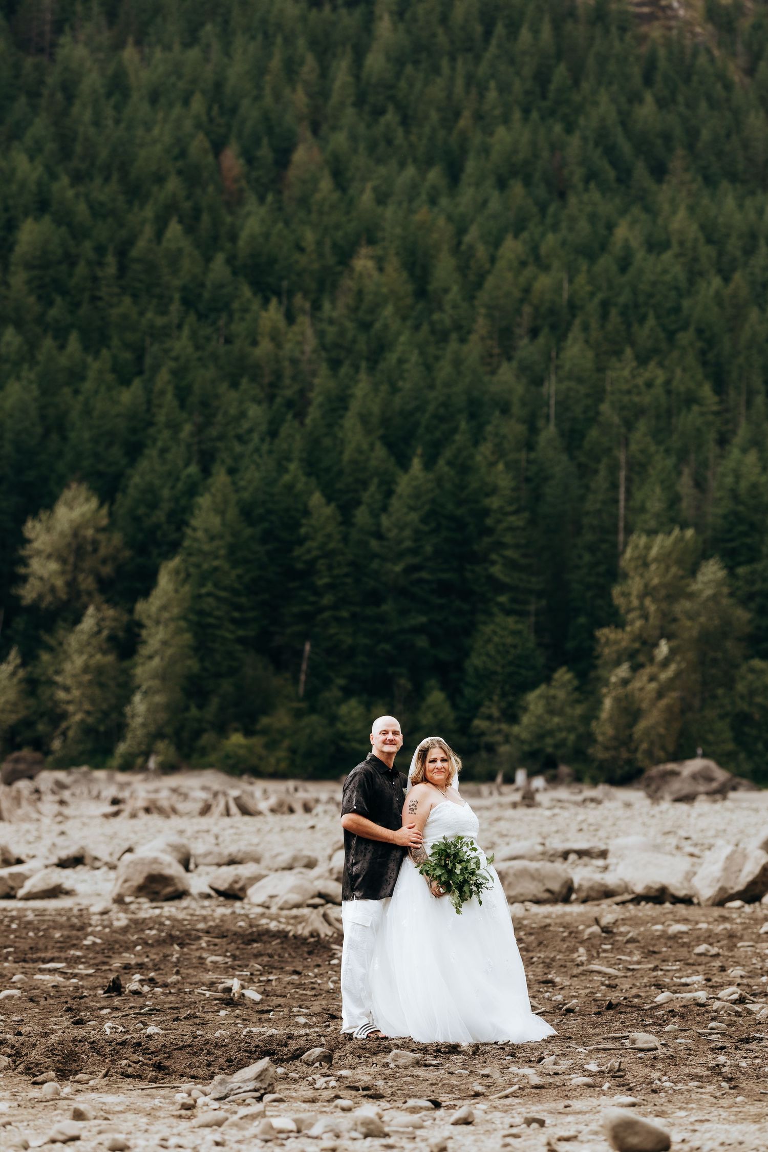 A couple in wedding attire sits on rocks with evergreen forest mountains in the background during their outdoor ceremony.