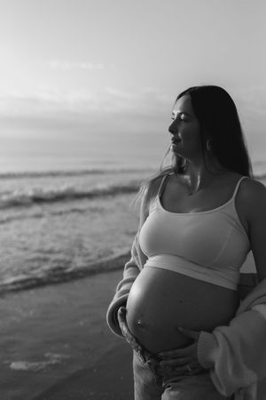 A series of black and white maternity photos showing a pregnant woman in a white top at the beach during sunset.