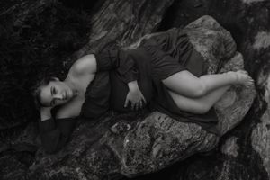 A person lies peacefully on a rocky surface while wearing a floral dress in a dramatic black and white photograph.