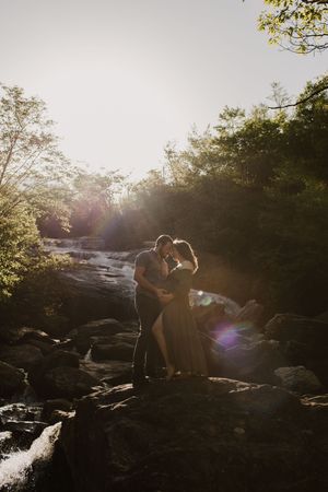 A silhouetted figure stands on rocky terrain near a stream during golden hour with sun flare in the background.