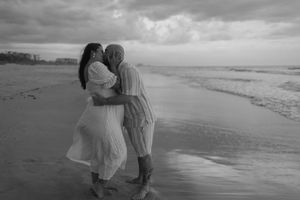 A romantic couple in white clothing shares tender moments along a misty beach shoreline at sunset.