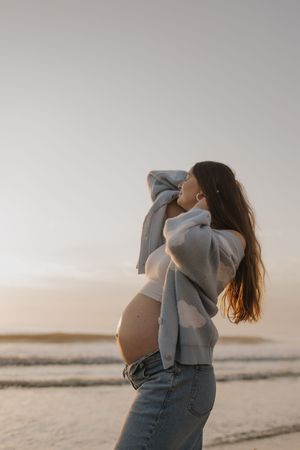 A pregnant woman in jeans and sweater stands on a beach at sunset with her hair flowing in the breeze.