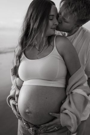 Intimate black and white maternity photo showing a couple embracing on a beach with the pregnant woman in a white tank top.