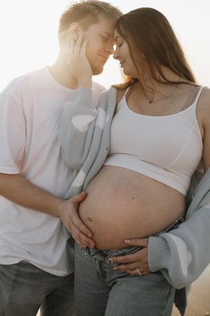 Intimate maternity photo of expecting couple embracing in soft sunlight, showing pregnant belly in casual attire.