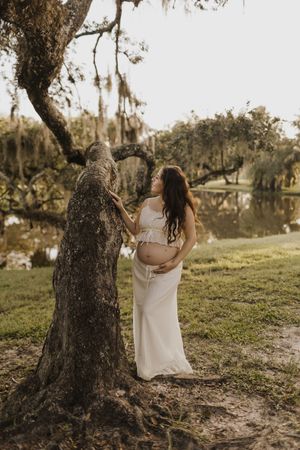 A pregnant woman in a white dress stands by a mossy tree near a serene lake at sunset.