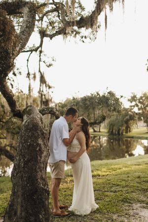 A romantic couple shares tender moments by a mossy tree near a serene lake at sunset.