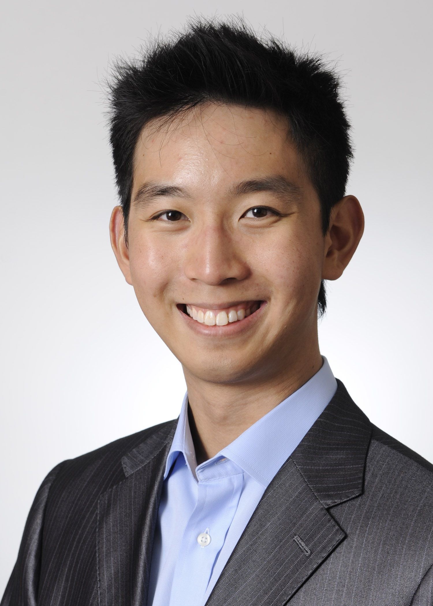 Professional headshot of someone in a gray suit and light blue dress shirt smiling against a white background.